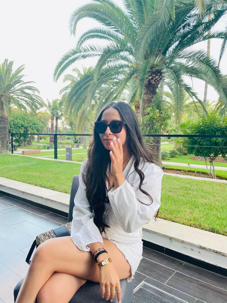 A woman in a white short-sleeved shirt dress and sunglasses sits on a patio chair, hand raised, with palm trees in the background.