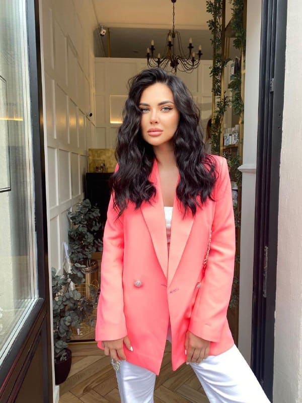 Anabelle stands in a doorway wearing a bright pink blazer over a white top and trousers, with a chandelier and plants behind her.