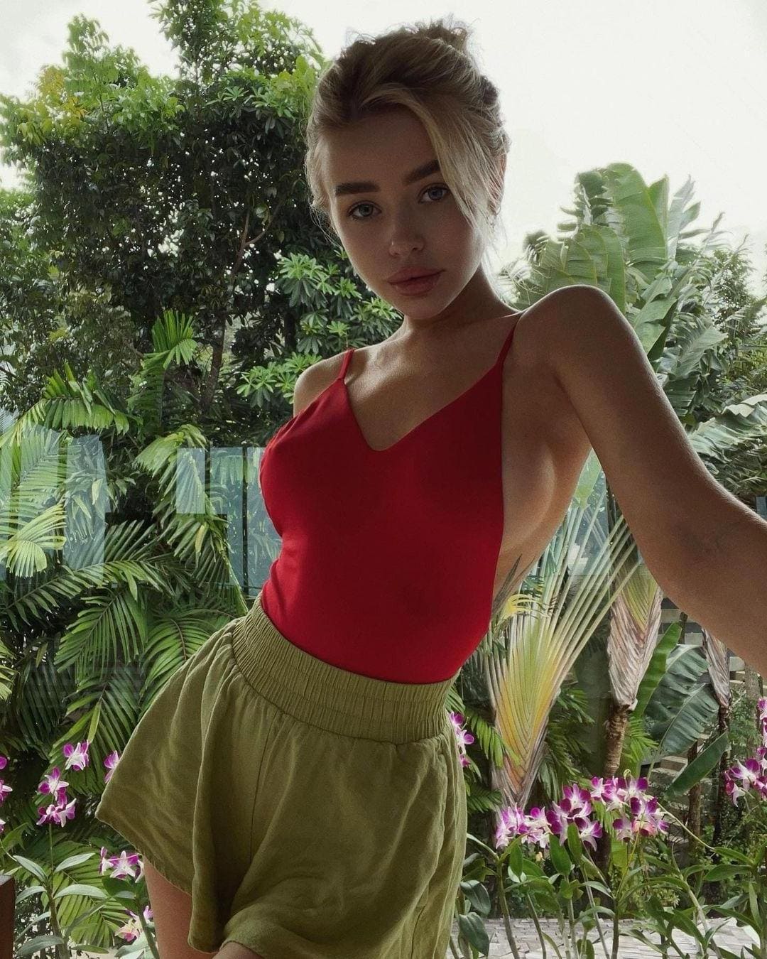 A woman with blonde hair in a loose updo wears a red spaghetti-strap top and olive shorts, standing before lush tropical foliage and purple flowers.