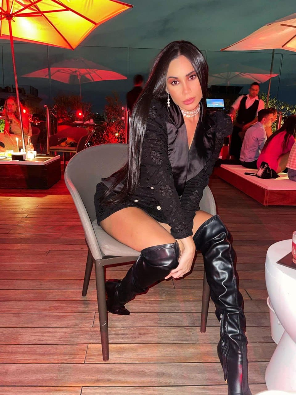 A woman in a black lace jacket, short skirt, and thigh-high boots sits on a grey chair at an outdoor evening venue with umbrellas behind.