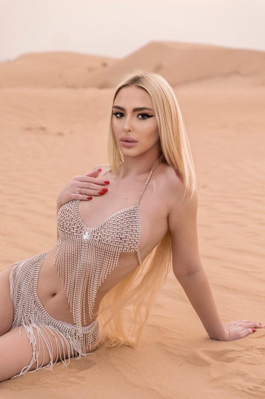 A blonde woman reclines on sand dunes wearing a beaded two-piece outfit with fringe, her right hand on her chest and left arm supporting her.