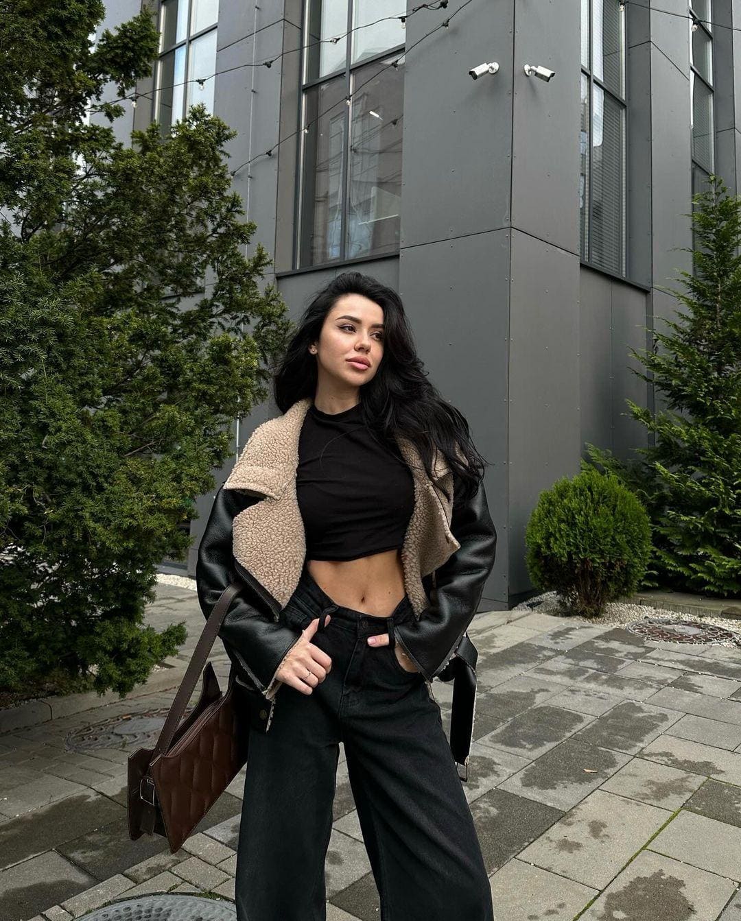 Nadine stands on a paved walkway before a modern building, wearing a black cropped top, wide-leg jeans, and a shearling-lined leather jacket.