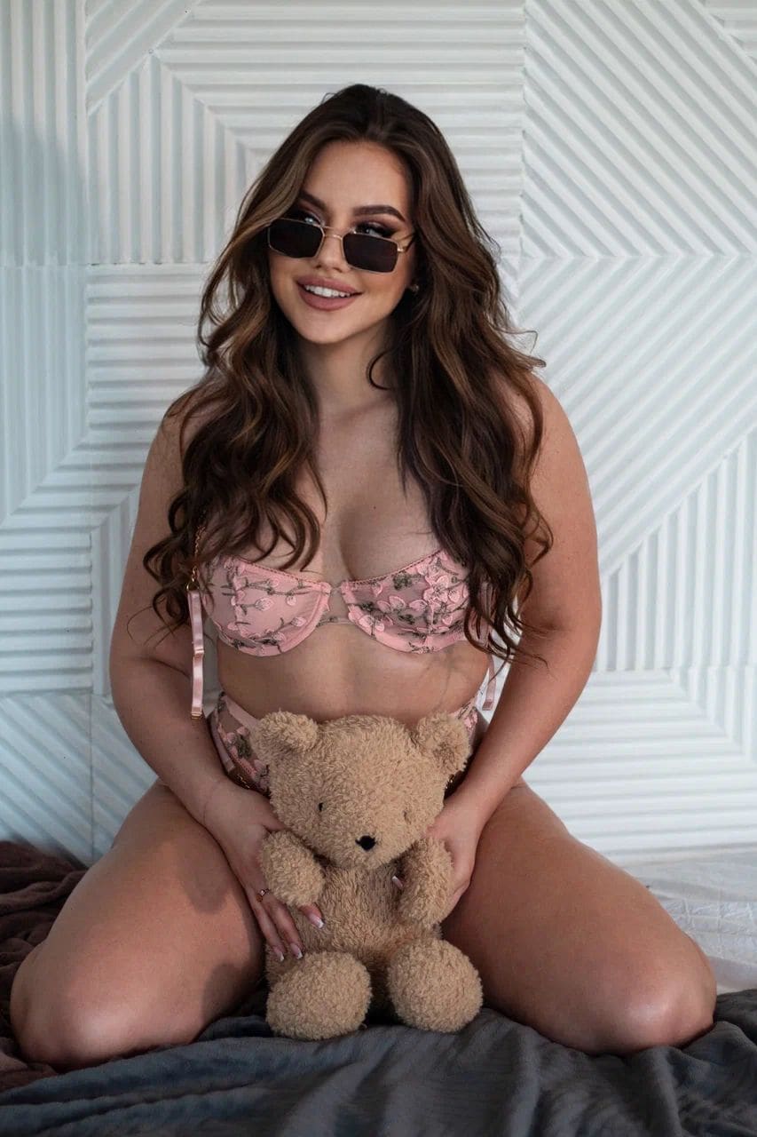 Woman in sunglasses and pink floral lingerie sits on a bed holding a teddy bear against a geometric wall.
