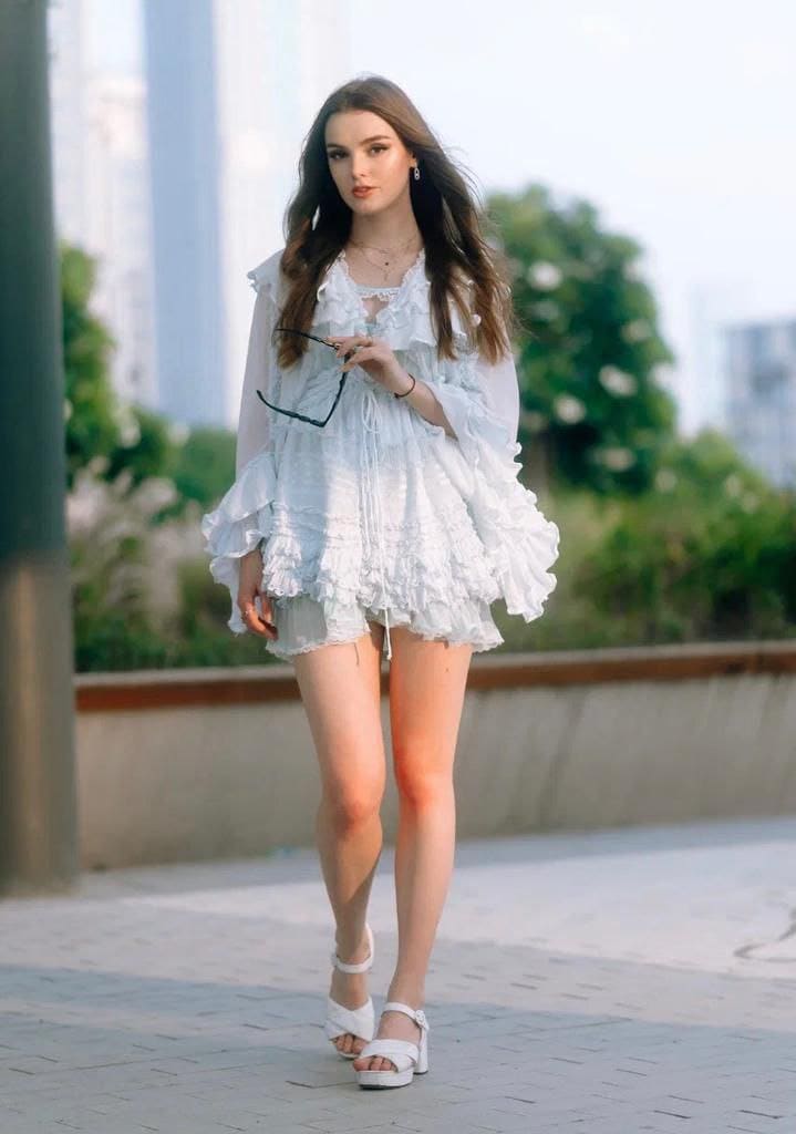 A woman in a white ruffled dress and platform sandals holds glasses on a paved walkway.