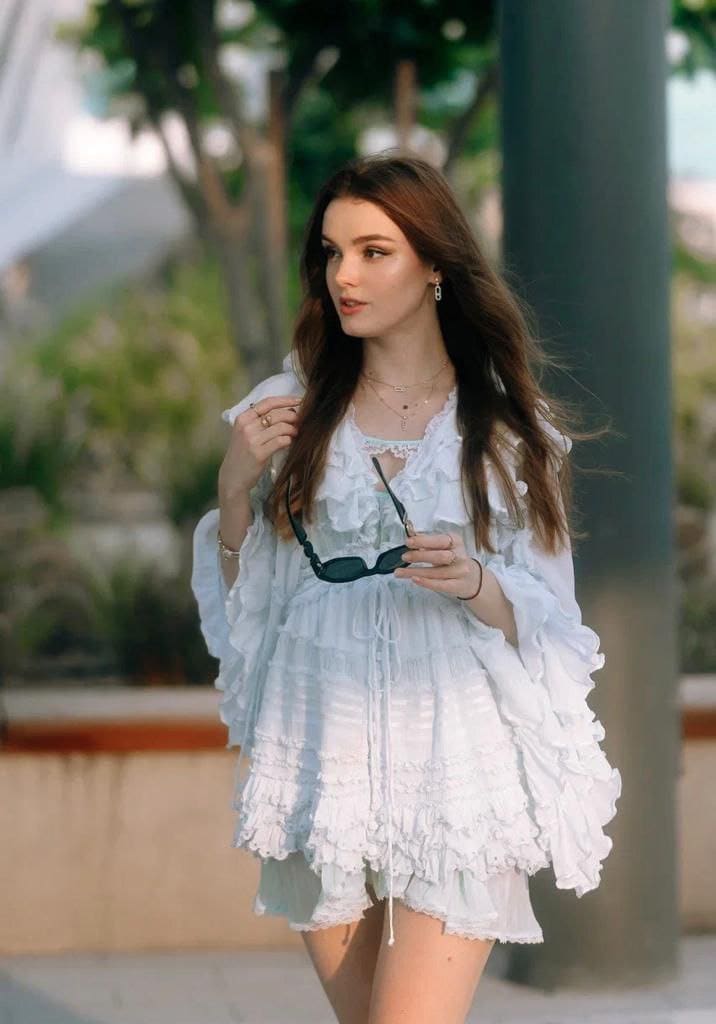 A woman in a white ruffled mini dress holds sunglasses, looking sideways near greenery.