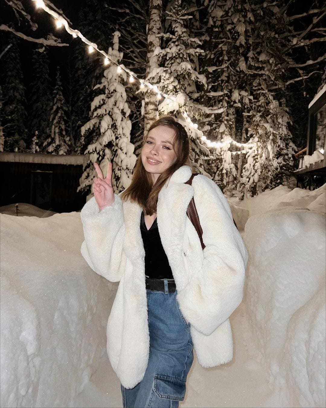 In a snowy night setting, she wears a white fluffy coat and jeans, making a peace sign under string lights.