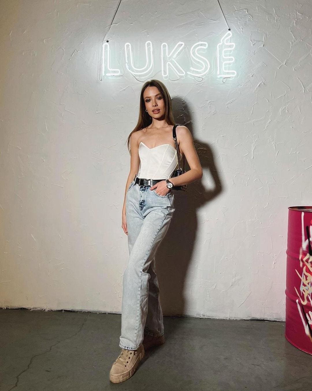 Zahra stands by a neon sign in a white strapless top, blue jeans, black belt, tan sneakers, and a shoulder bag.