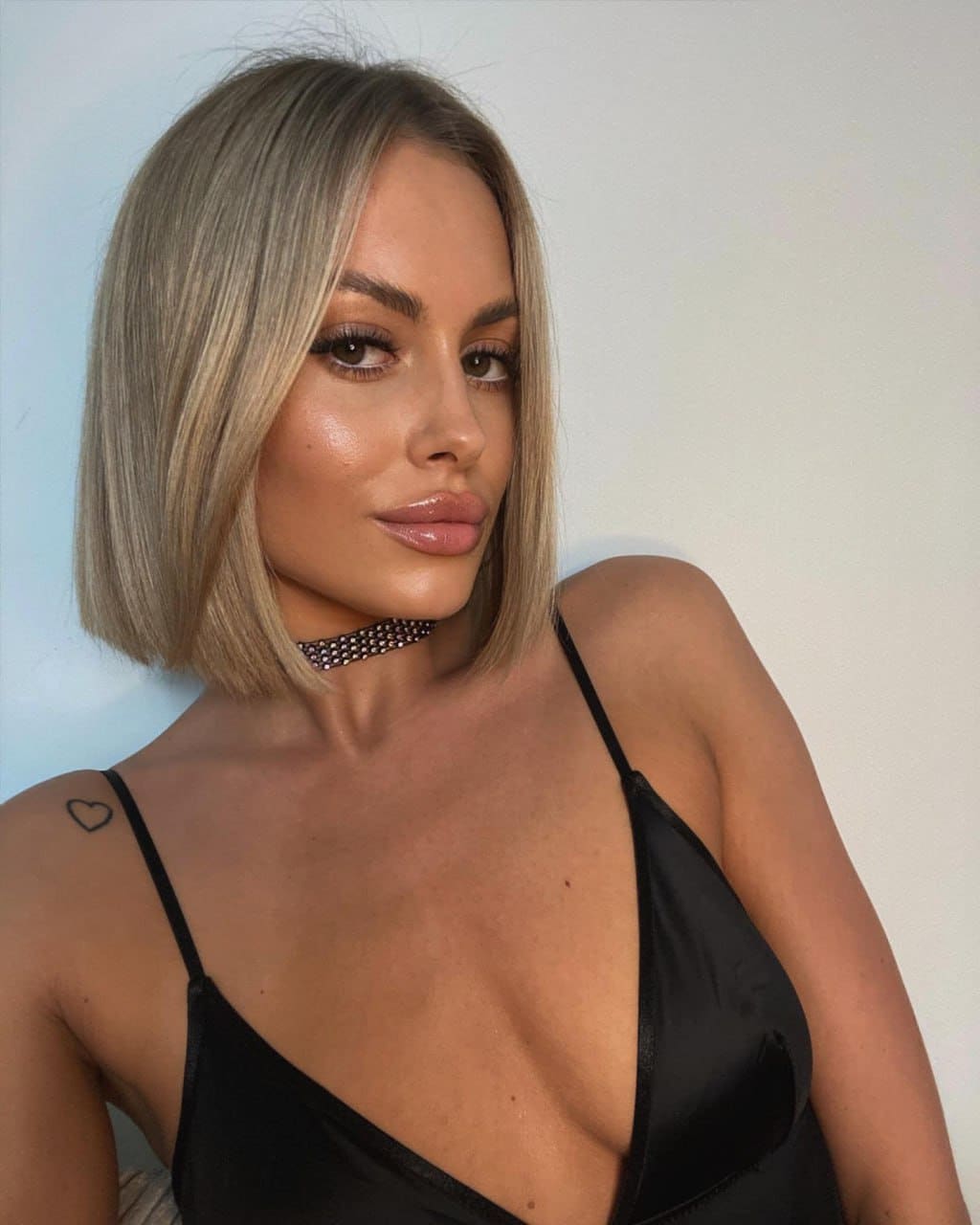 Viola with a blonde bob wears a black satin slip top and studded choker, posing against a plain light wall.