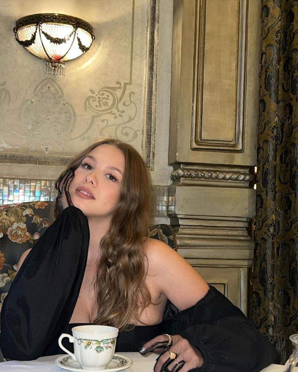 A woman in a black off-the-shoulder top sits at a table in an ornate room, hand near her chin, with a teacup.