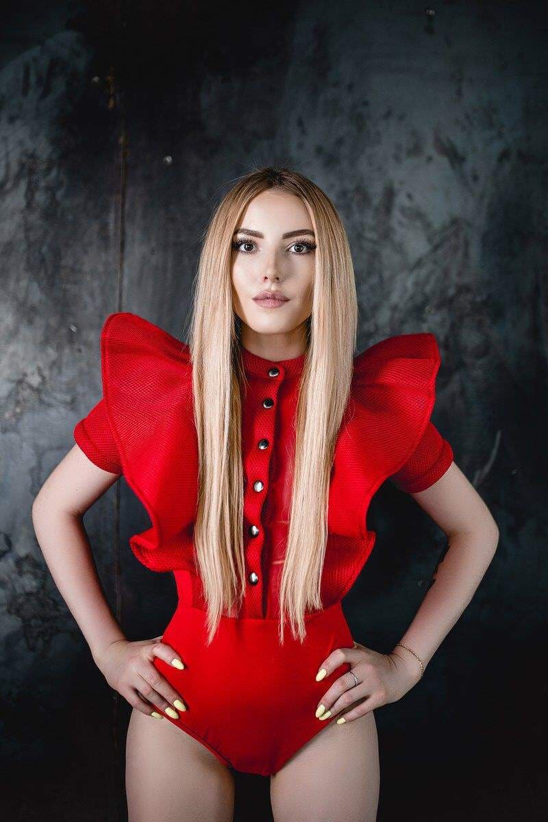 A woman stands with hands on hips in a red one-piece with ruffled shoulders and black buttons against a dark backdrop.