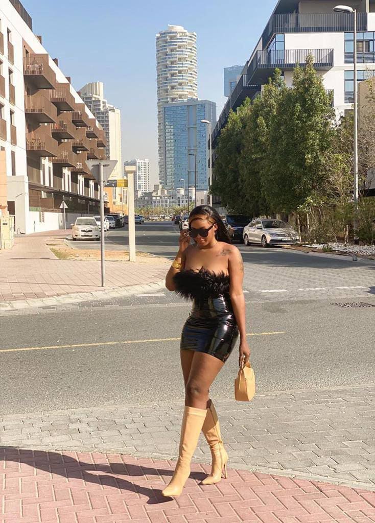 A woman stands on a city sidewalk in a strapless black feathered top, shiny skirt, and tan knee-high boots, holding a small handbag.