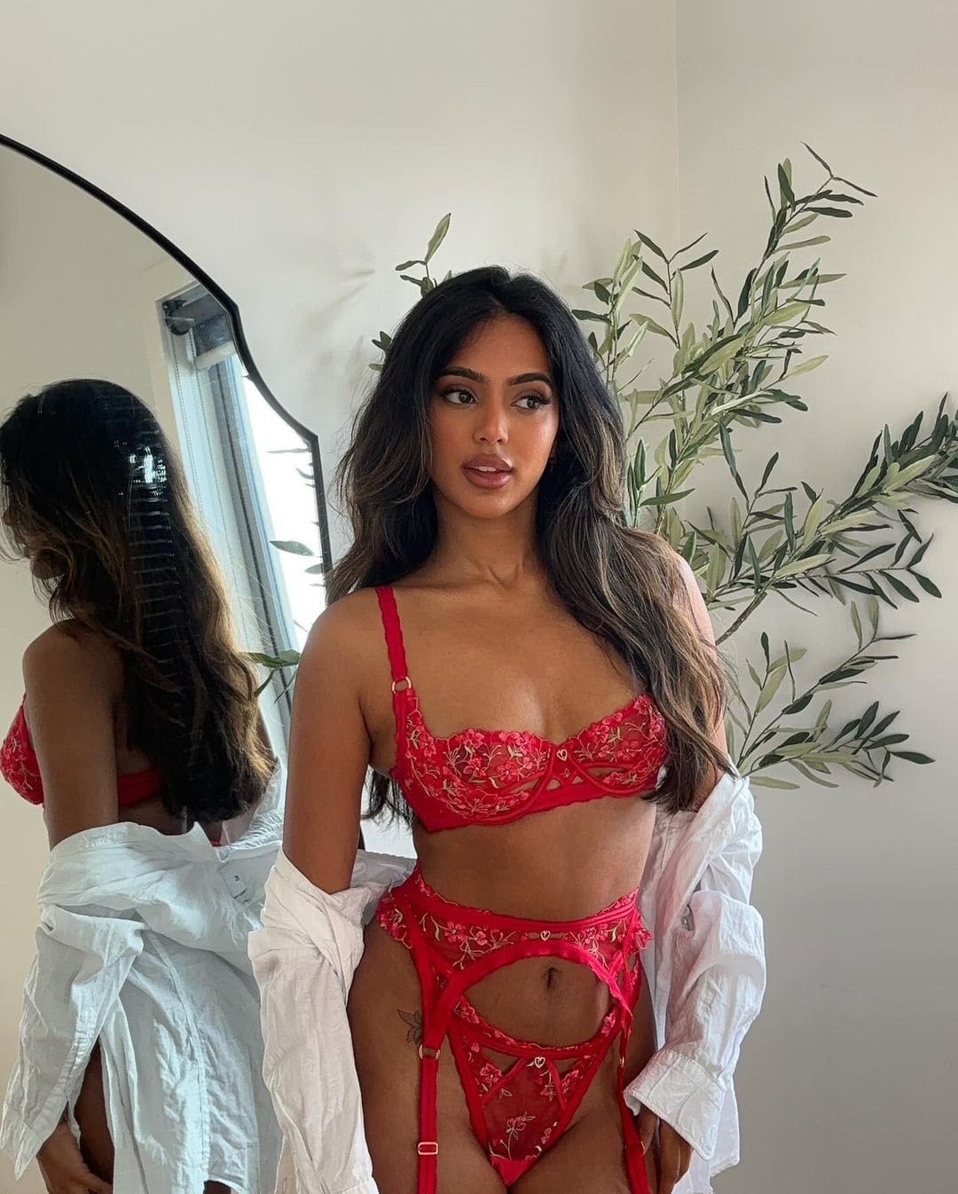 Standing indoors, she wears a red lace set with garters and an unbuttoned white shirt off-shoulder, near a plant and mirror.