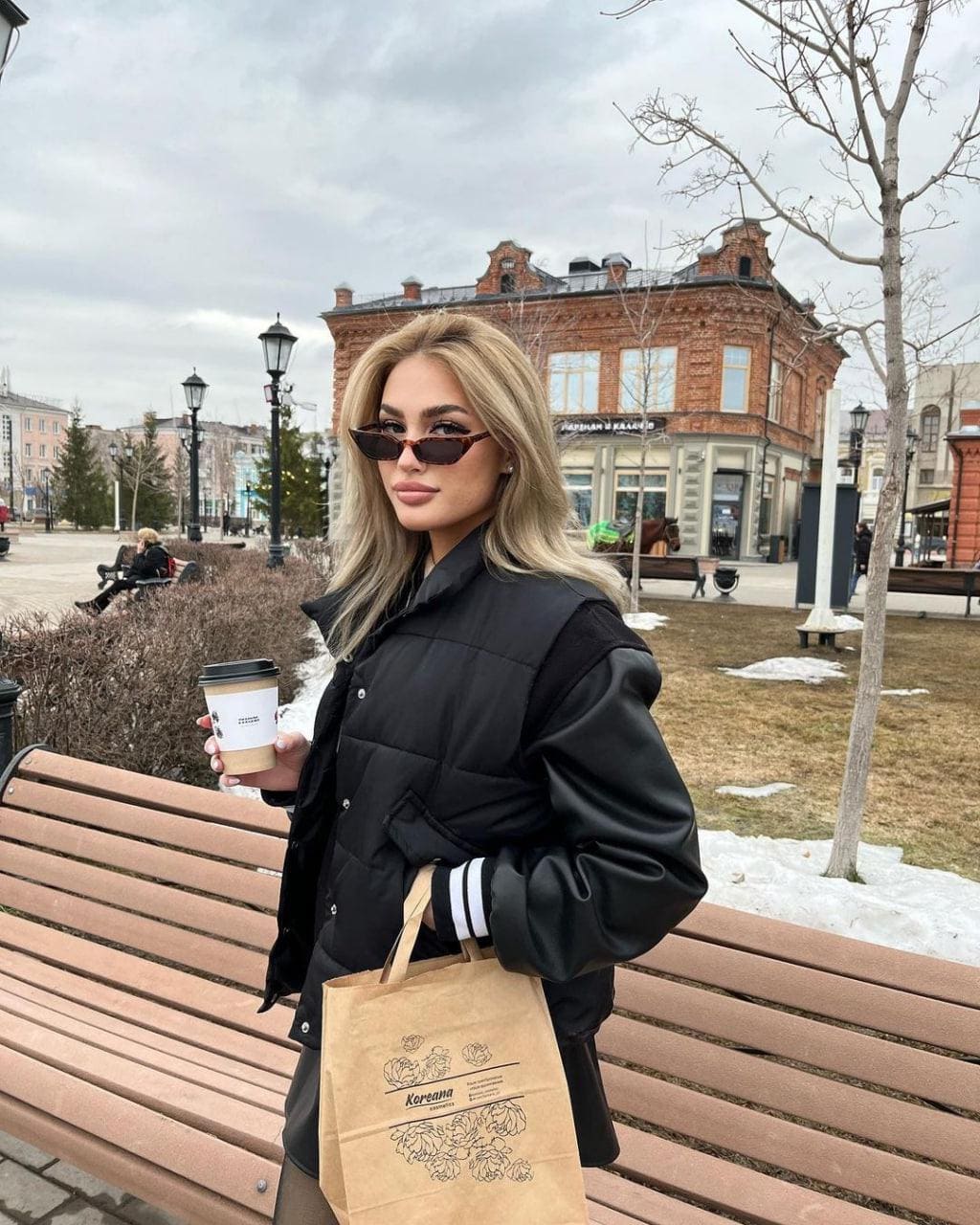 Standing in a park, a woman in a quilted jacket and sunglasses holds a coffee cup and paper bag near a bench.