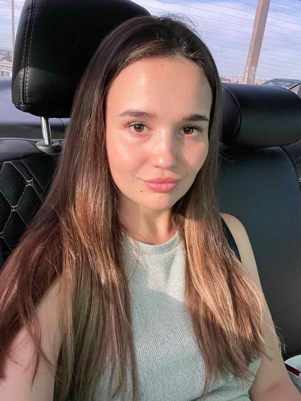 Ksenia sits in a car back seat wearing a light grey sleeveless top, with a black leather headrest and window behind her.