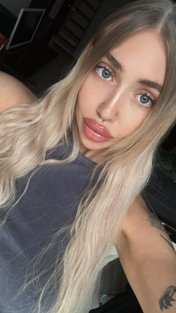 Close-up indoor selfie features a woman with wavy blonde hair and blue eyes in a dark grey sleeveless top, against a dark background.