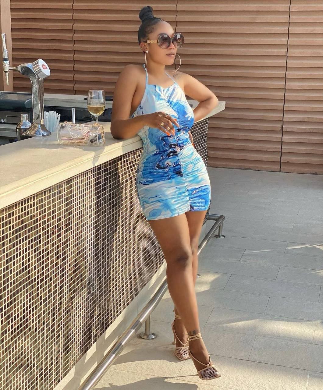 A woman in sunglasses leans on a tiled bar counter in a blue-and-white patterned halter dress, with a glass of white wine beside her.