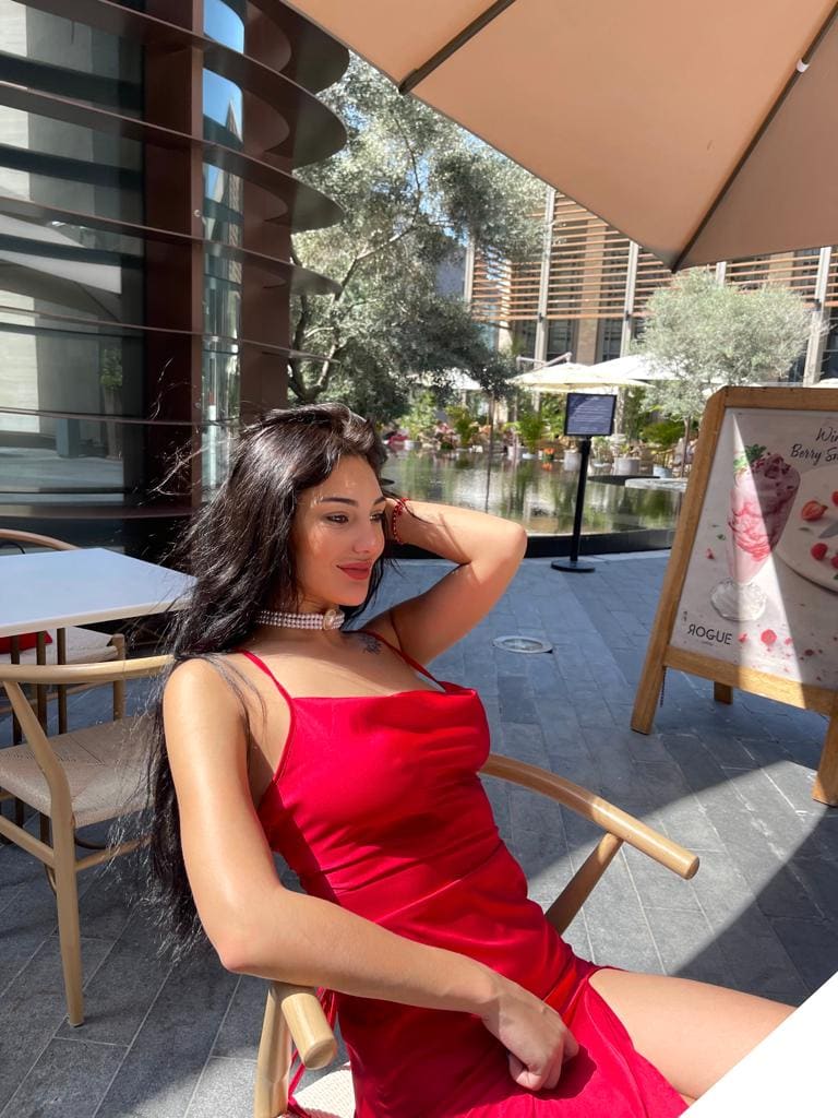 A woman in a red slip dress and pearl choker sits in a wooden chair on a patio, hand behind head, near a menu board.