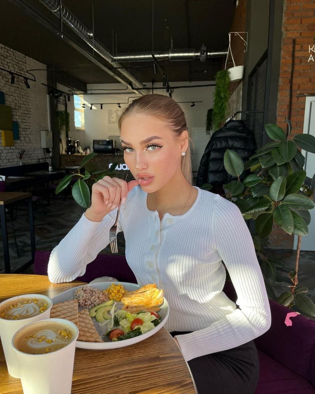 Mila sits at a cafe table in a white ribbed top, holding a fork near her mouth with food and coffee in front.