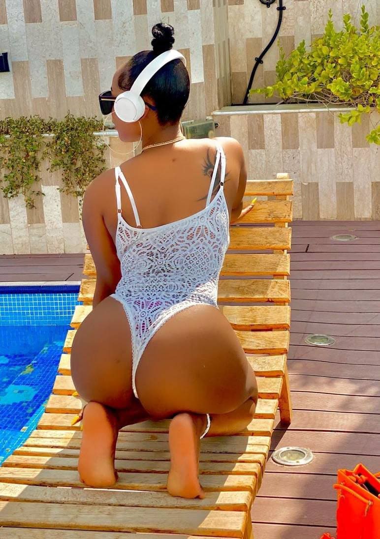 Viewed from behind, a woman in a white lace swimsuit and headphones kneels on a poolside lounger facing a tiled wall and greenery.