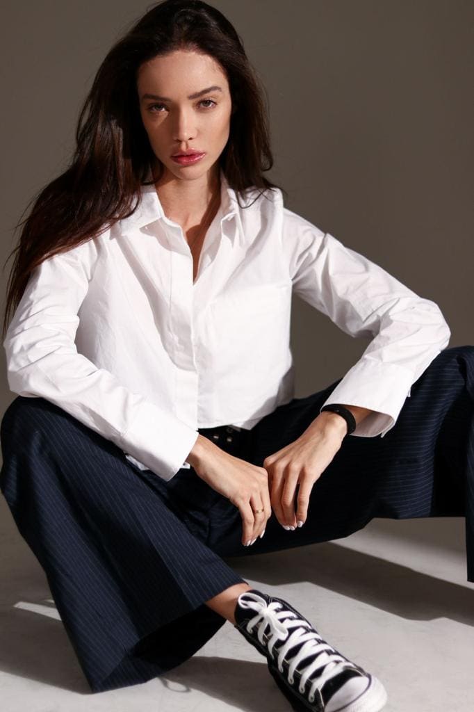 Angela sits on a light floor against a gray wall, wearing an open white shirt, pinstriped trousers, and black sneakers.