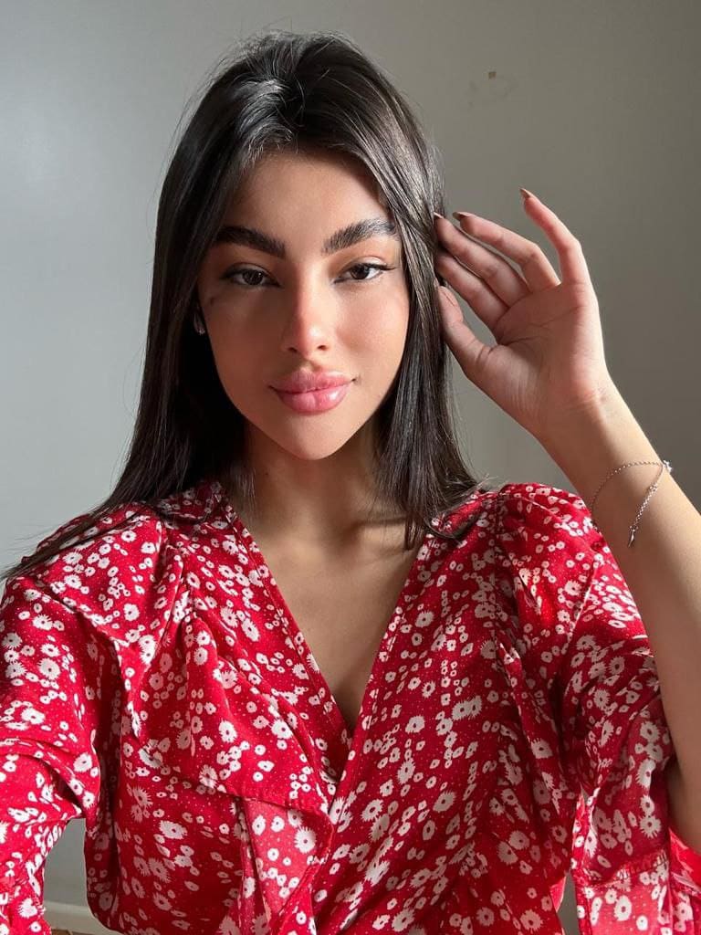CHLOE poses in a red floral V-neck blouse, touching her long dark hair against a plain gray wall.