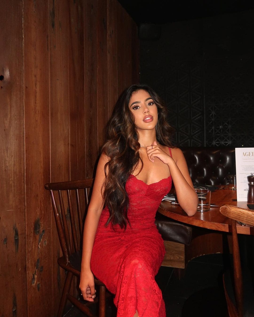 Seated in a dim, wood-paneled room, she wears a red lace dress with thin straps, hand near chin, beside a table with a glass.