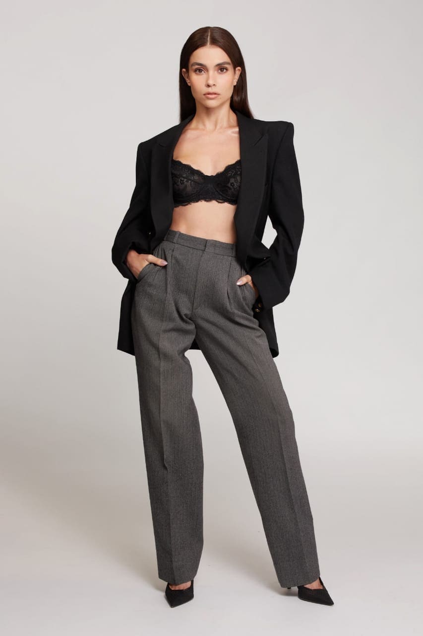 A woman faces forward with hands in the pockets of grey pinstripe trousers, wearing an open black blazer over a lace bra and heels.