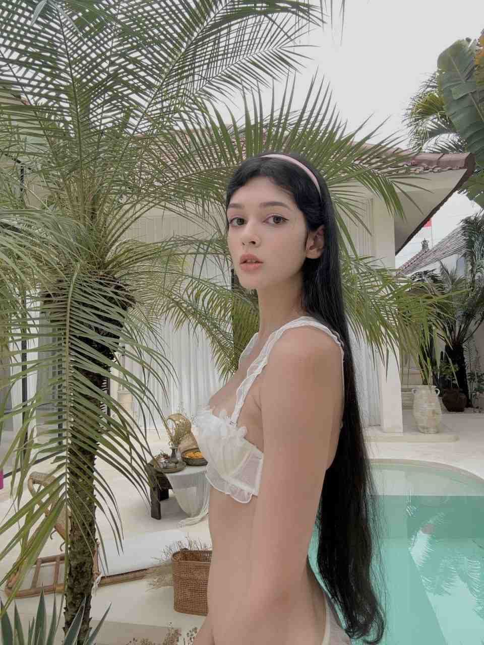 Profile view of a woman in white lace lingerie and a headband standing by a pool, with palm fronds and a white building in the background.