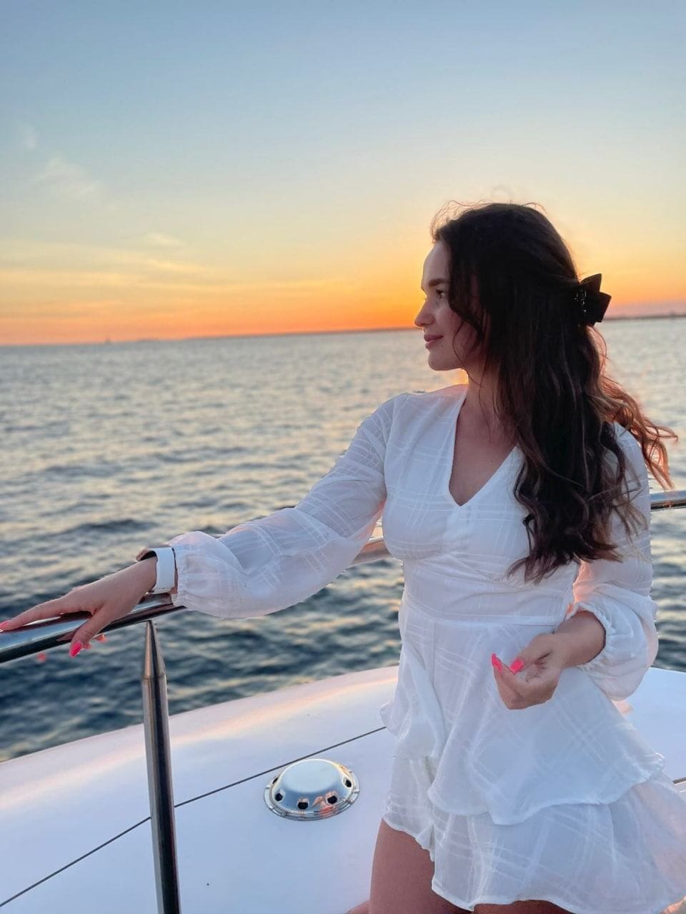 A woman in a white ruffled dress stands on a boat deck at sunset, resting one hand on the metal railing with her hair tied back.