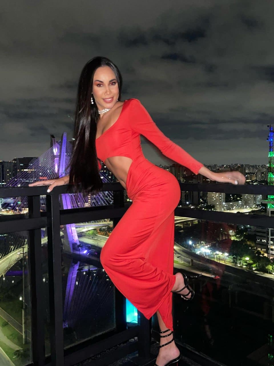 A woman stands on a rooftop balcony at night in a red dress with a side cutout, leaning on the railing against glowing city lights.